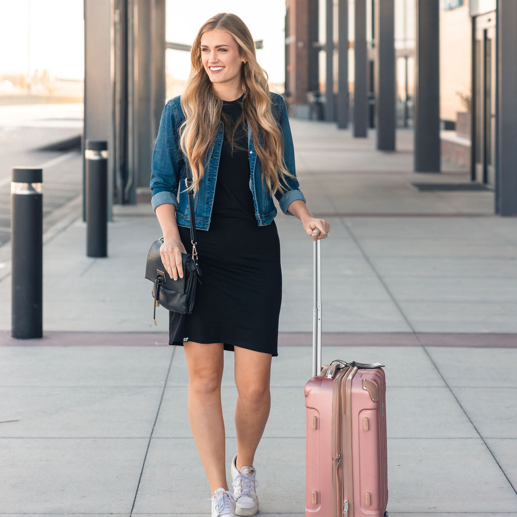 A woman styled in the Everyday Merino Dress in black, complemented by a denim jacket and carrying a pink suitcase